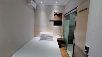 A narrow hotel room with a bed, a small table, and a glass door leading to a bathroom.