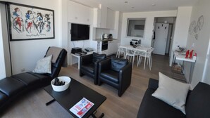 Ein modernes Wohnzimmer mit einer Couch, einem Couchtisch und einem Fernseher. Es gibt eine Essecke mit Tisch und Stühlen. Im Hintergrund ist die Küche zu sehen.