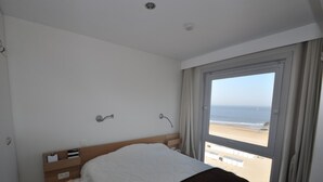 Ein Schlafzimmer mit Bett, Nachttisch, Fenster mit Blick auf den Strand und Deckenleuchte.