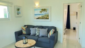 A living room with a grey sofa, a round wooden coffee table, and two wall-mounted paintings depicting tropical scenes. - Luxurious 1-bedroom condo in Sosúa with AC, WiFi, Backyard, Terrace, and Balcony (Sosúa)