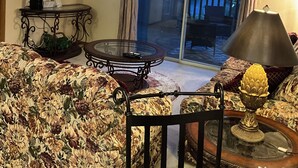 A living room with a ceiling fan, a television, a glass table, and a sofa with floral upholstery. - Cozy 2-bedroom condo- right beside SCD (Branson)