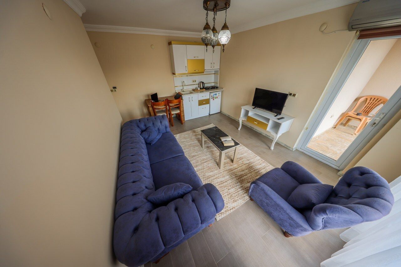 A living room with a sofa and armchairs, a small table, a TV, and a kitchen in the background.