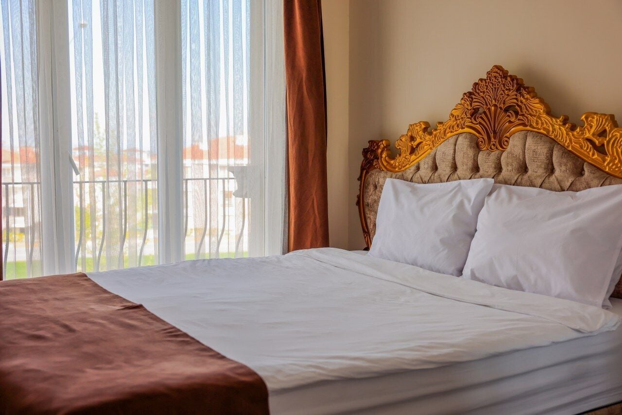A neatly made bed with a brown headboard, white bedding, and a view of a balcony through sheer curtains.