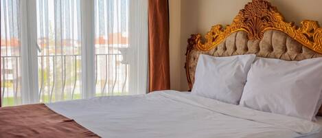 A neatly made bed with a brown headboard, white bedding, and a view of a balcony through sheer curtains.