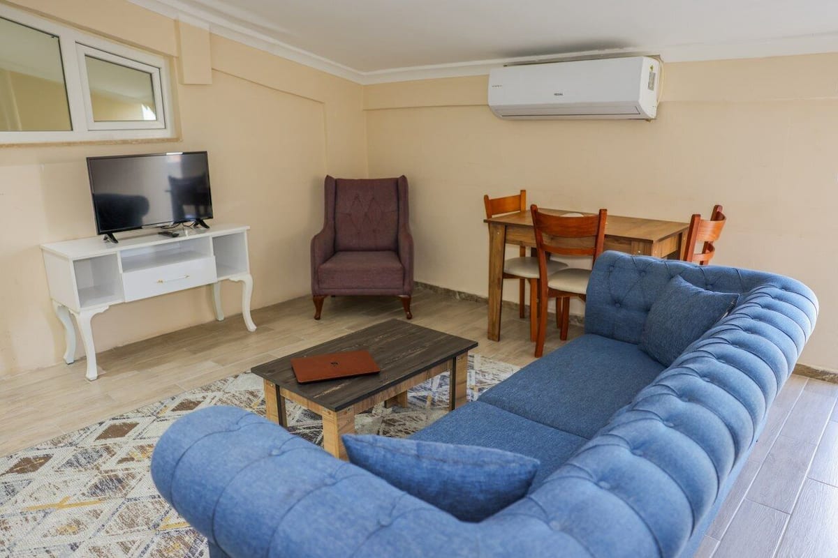 Ein Wohnzimmer mit blauem Sofa, einem Holztisch, einem Fernseher auf einem weißen Schrank, einem violetten Sessel und einem Holzessttisch mit Stühlen.
