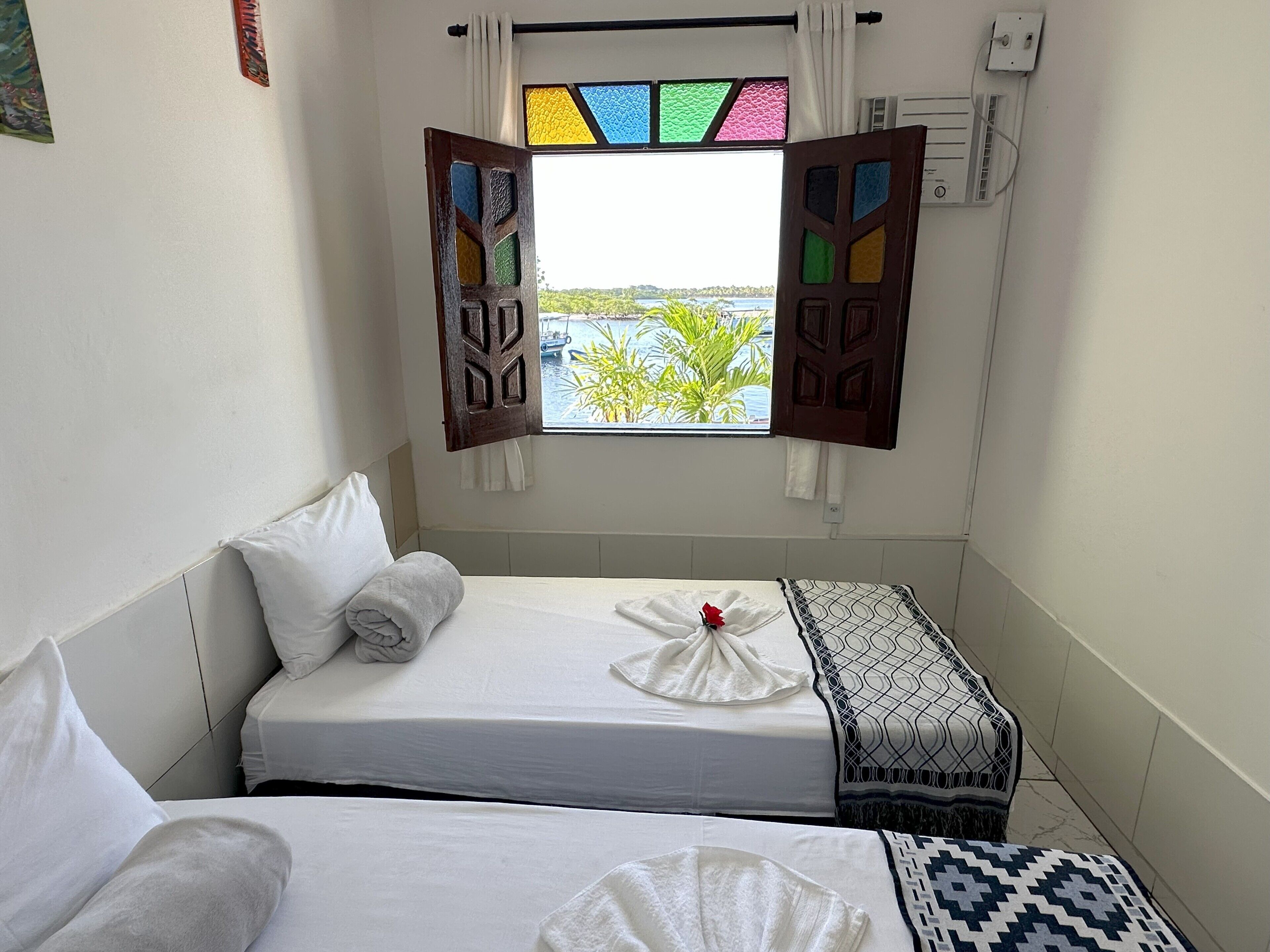 A small, clean room with two beds, a window with colorful shutters, and a view of a body of water.