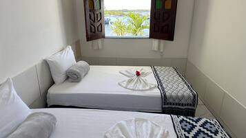 Quarto pequeno e limpo, com duas camas, janela com persianas coloridas e vista para um corpo d'água.