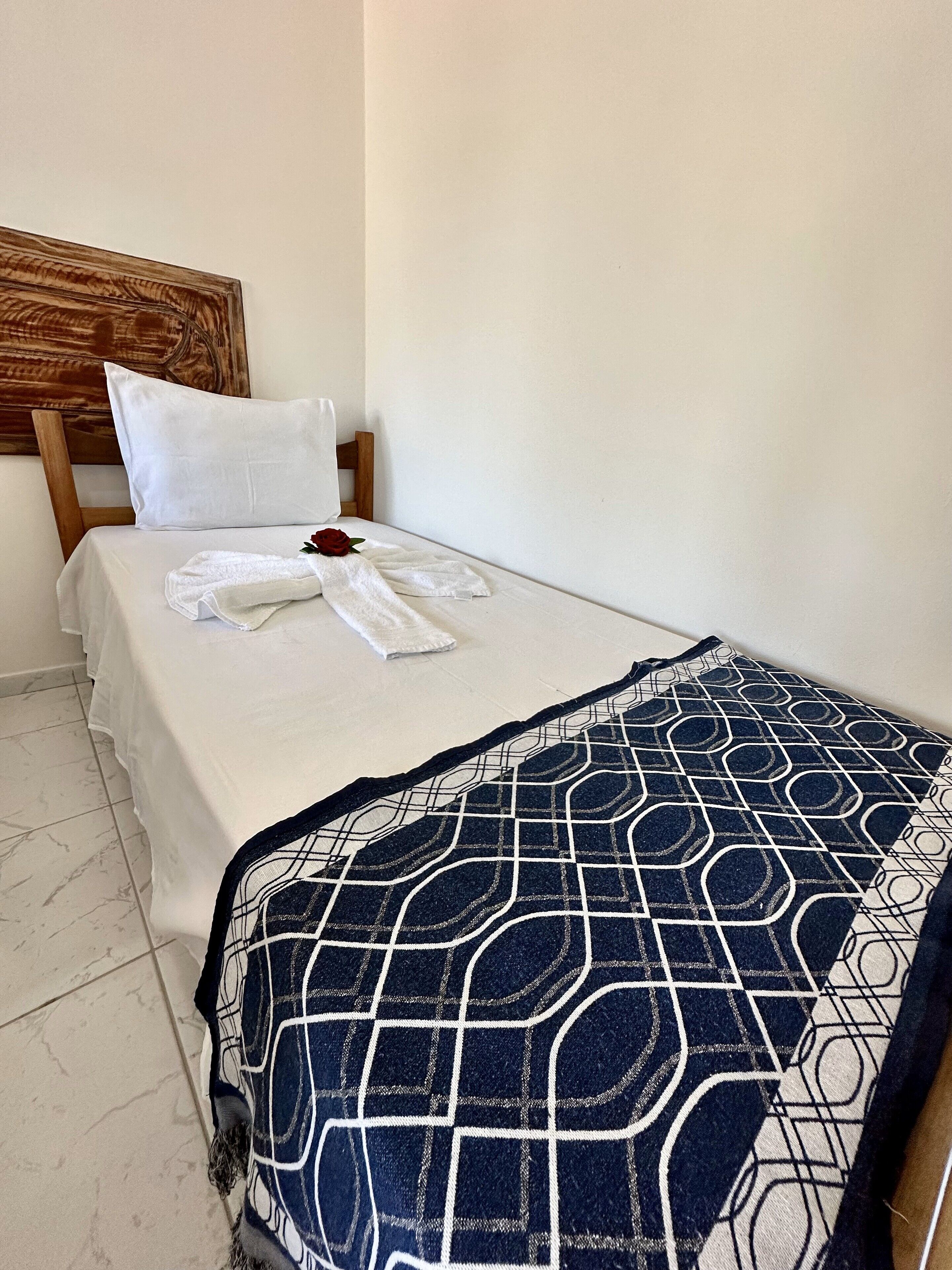 A neatly made bed with a white sheet, a white pillow, and a single red rose. A blue and white patterned blanket is draped over the bed.