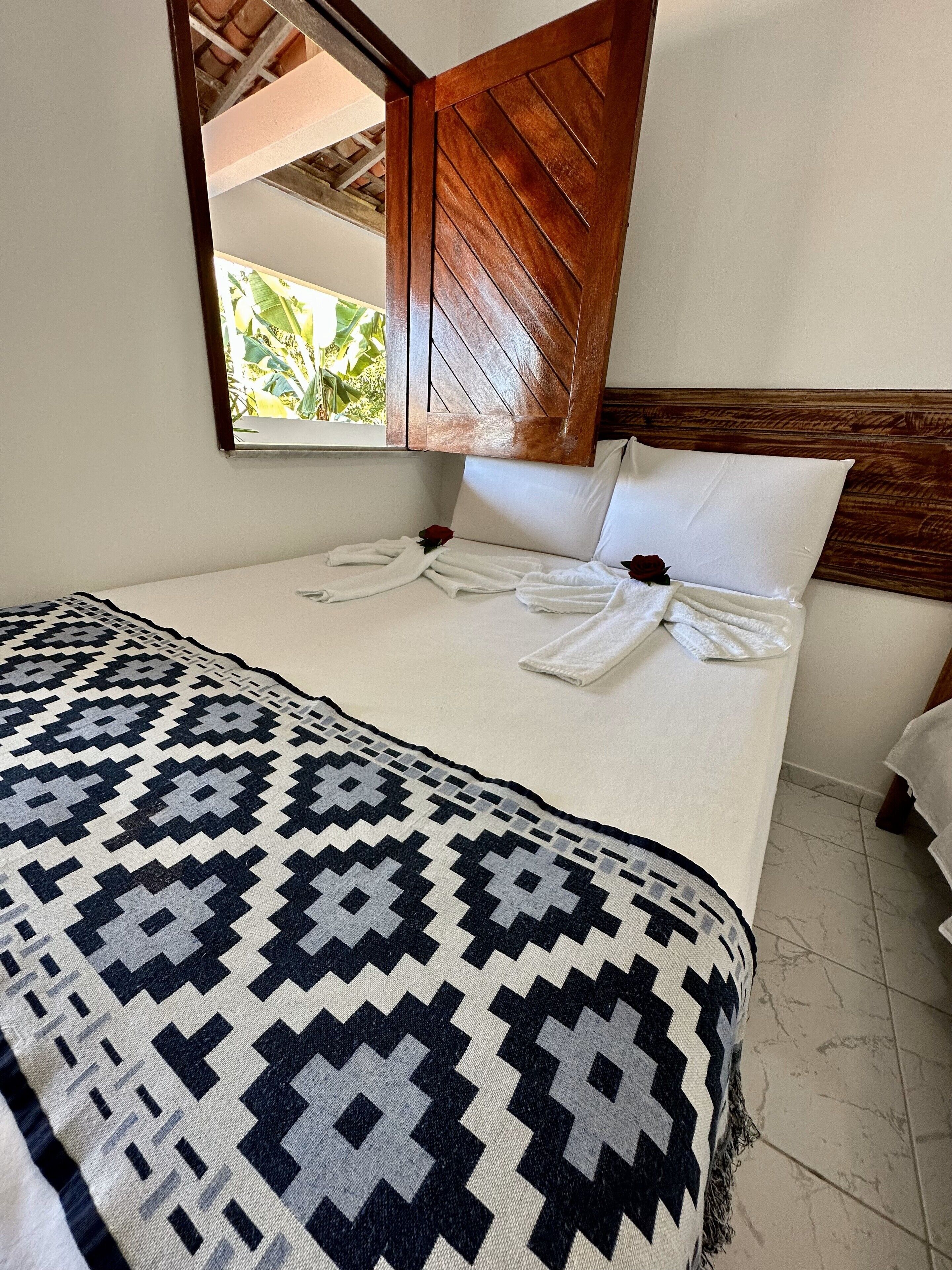Una cama bien hecha con un edredón estampado, cabecera de madera y ventana con vistas a zonas verdes.