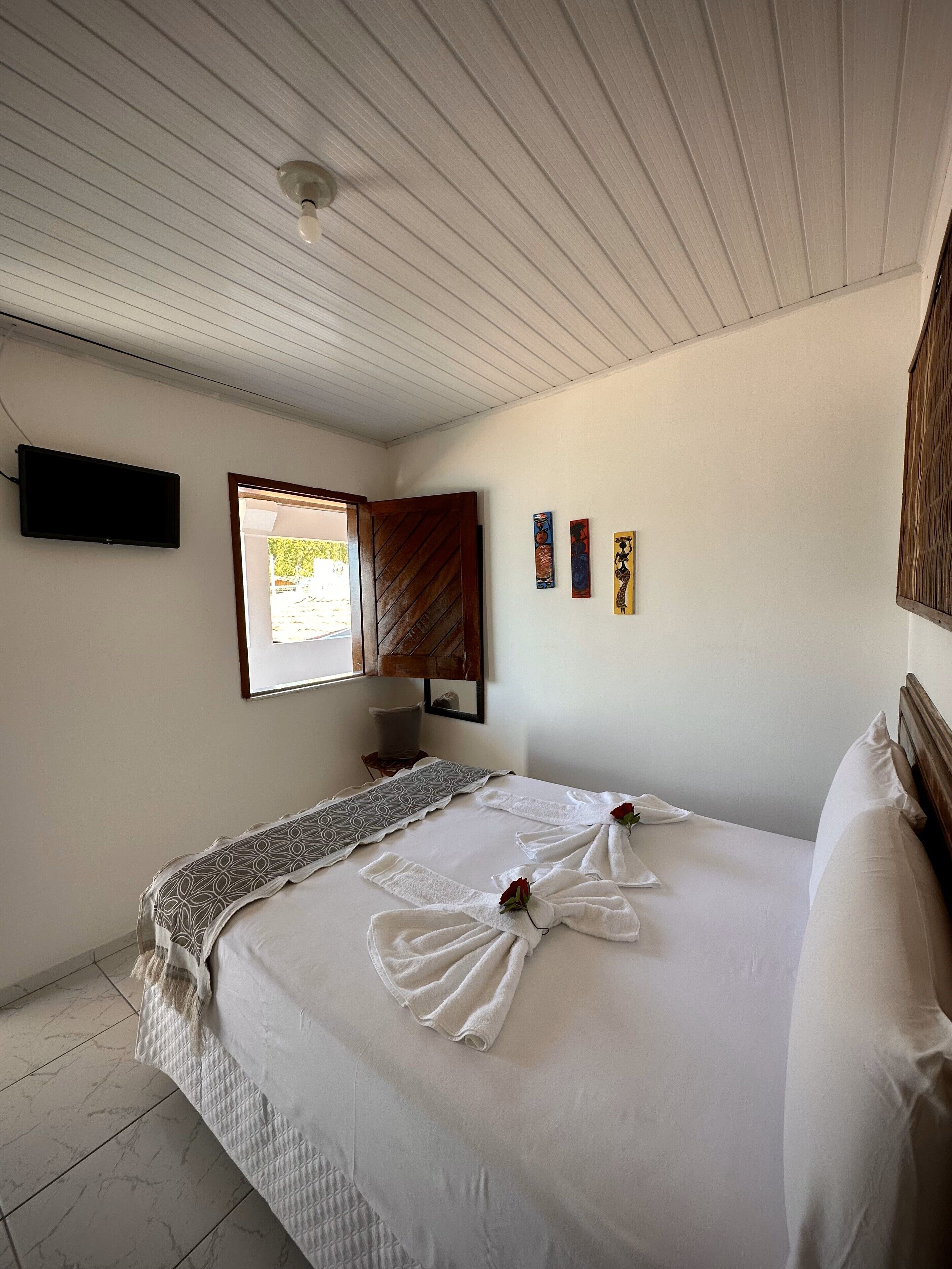 Un dormitorio con cama, televisor de pantalla plana montado en la pared, ventana con vistas y tres cuadros colgados.