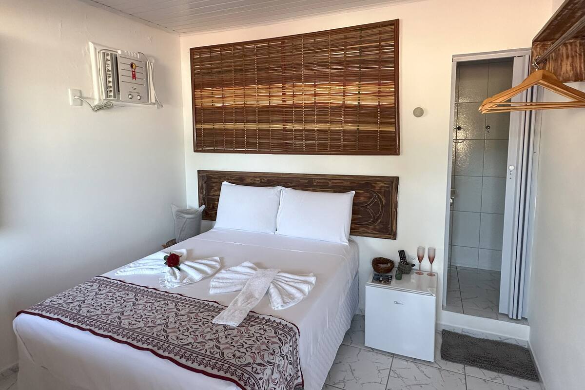 Ein ordentlich bezogenes Bett mit weißen Leinentextilien, einem Holzkopfteil und einer dekorativen Tagesdecke. Ein kleiner Nachttisch bietet Platz für eine Fernbedienung, eine Schale und zwei Gläser. Über dem Bett ist ein Sichtschutz aus Holz angebracht, und zur Rechten ist eine Duschkabine zu sehen.