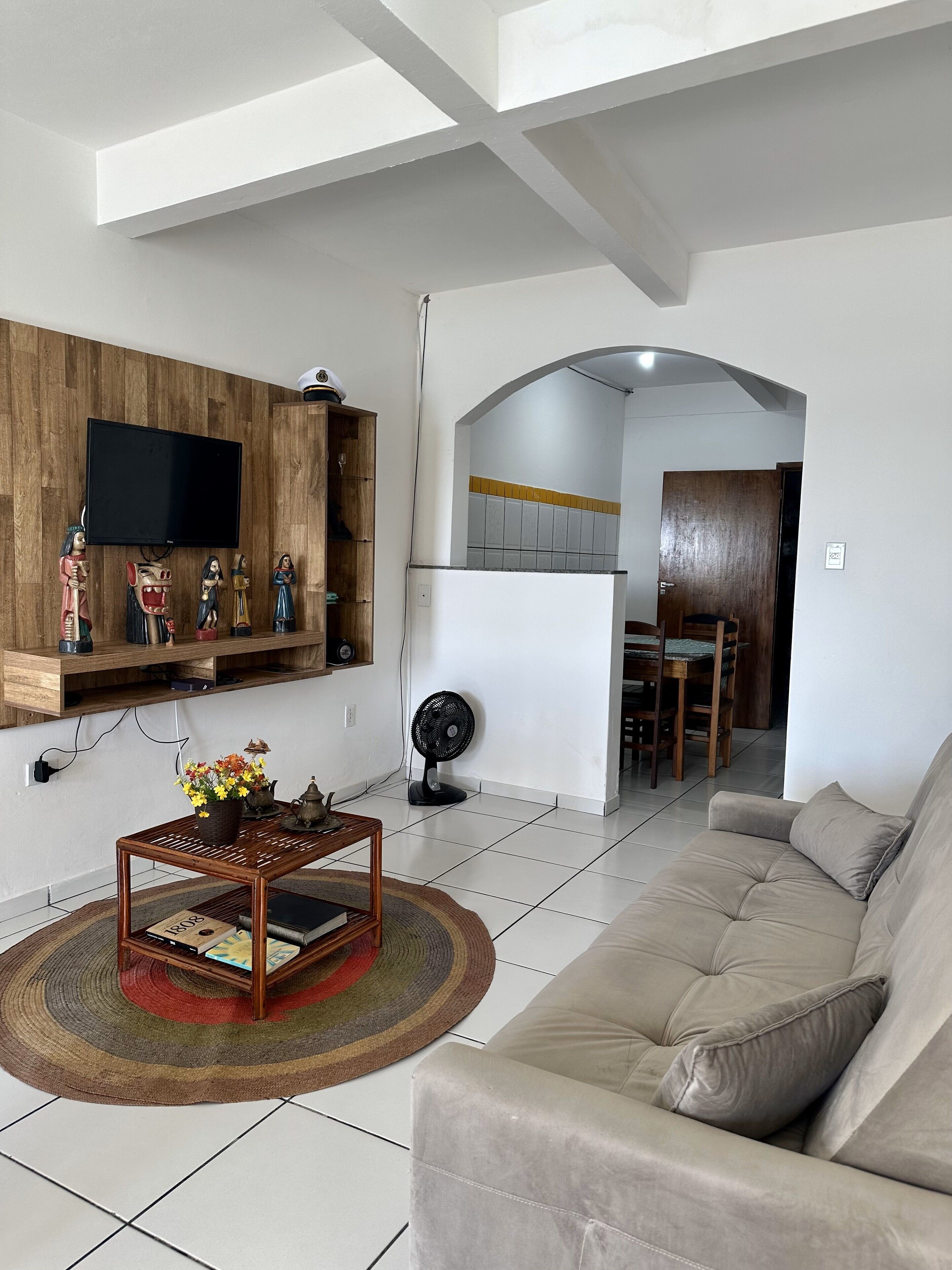 Una sala de estar con un sofá, una mesa de centro con libros, un televisor fijado en una pared de madera y un ventilador en el suelo.