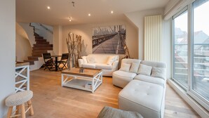 A modern living room with a staircase, a white sofa, a coffee table, and a painting of a bridge. - Duplex in Knokke-heist Near the Beach (Knokke-Heist)