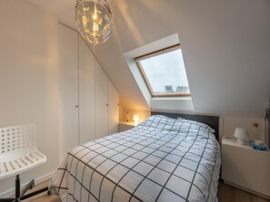 A bedroom with a skylight, a bed with a checkered duvet, a white chair, and a bedside table with a lamp. - Duplex in Knokke-heist Near the Beach (Knokke-Heist)
