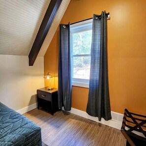 A room with a sloped ceiling, a window with dark curtains, a bedside lamp, a wooden nightstand, and a blue quilted bed.