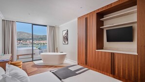 A modern hotel room with a large bed, a bathtub, a TV, and a balcony with a city view.