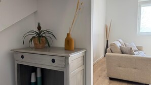 A room with a light-colored sofa, a small wooden side table with a plant and a vase, and a window with natural light. - Village house in the countryside, T3 duplex with master suite. (Ville-sous-Anjou)