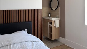 A modern hotel room with a bed, wooden panel headboard, a bathroom with a sink and mirror, and a window providing natural light. - Village house in the countryside, T3 duplex with master suite. (Ville-sous-Anjou)