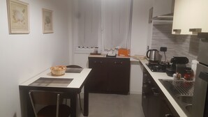 A compact kitchen with a dining table, chairs, and a small island. There are framed pictures on the wall, a window with curtains, and a coffee machine on the counter. - MILAN NAVIGLI LARGE APARTMENT VIA SAVONA-TORTONA (Milano)