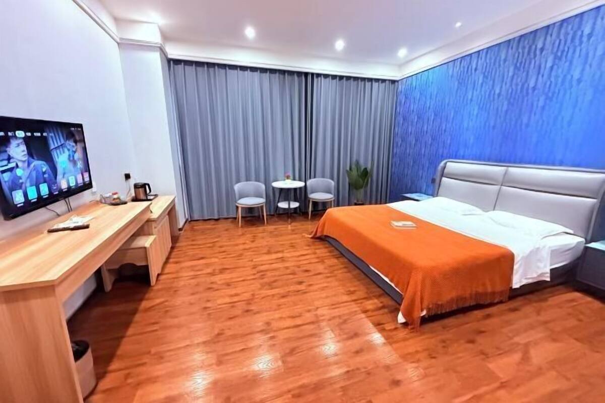 Ein Hotelzimmer mit Holzboden, einem Bett mit orangefarbener Bettdecke, einem Schreibtisch mit Fernseher, zwei Stühlen und einer blauen Wand mit Struktur.