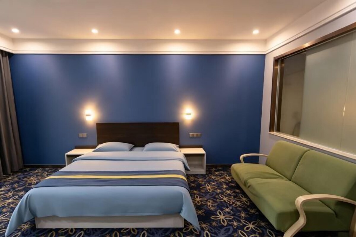 A hotel room with a large bed, two bedside tables, a green sofa, and a large window.