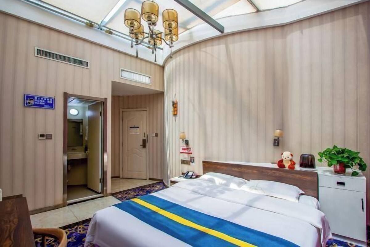 Ein Hotelzimmer mit Bett, Schreibtisch, Stuhl, Nachttisch, Lampe, Teddybär und Topfpflanze.