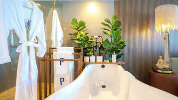 A bathroom with a bathtub, a towel rack with robes, a plant, and a lamp.