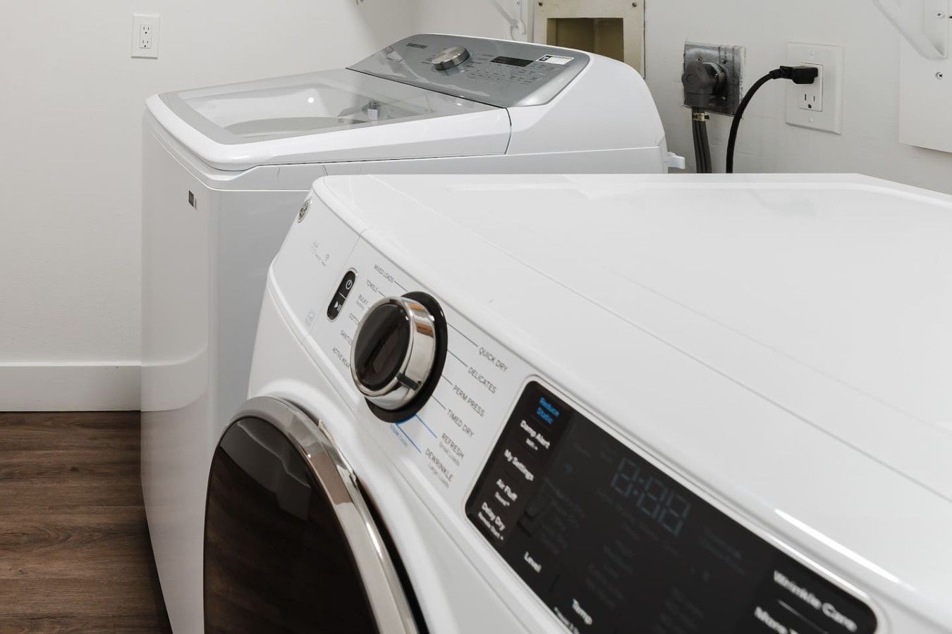 A front view of a modern, white, front-loading washing machine with a digital display, a control panel, and a power cord plugged into a wall outlet.
