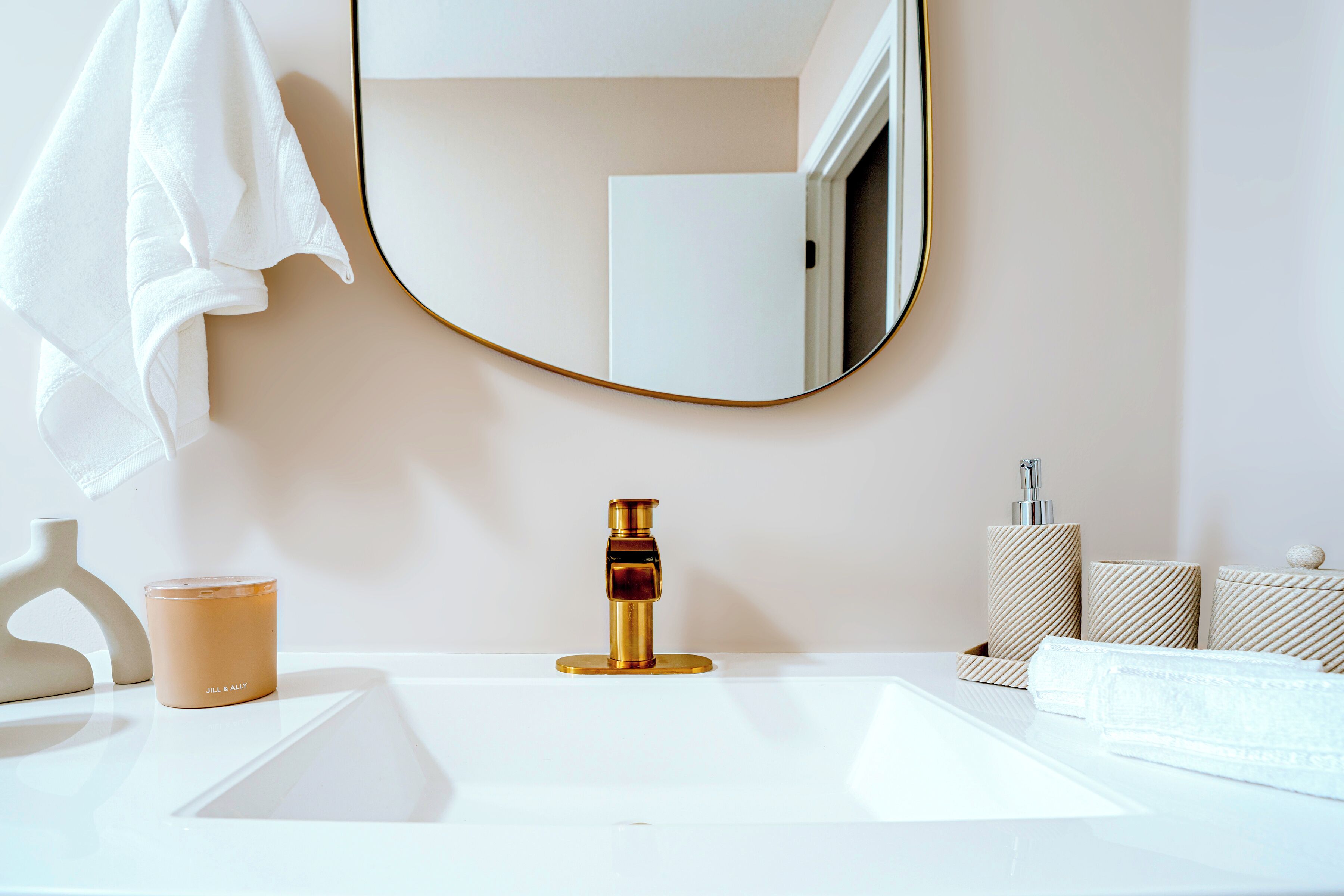 Un bagno con un lavandino bianco, un rubinetto dorato, uno specchio rotondo, un asciugamano bianco e una candela.