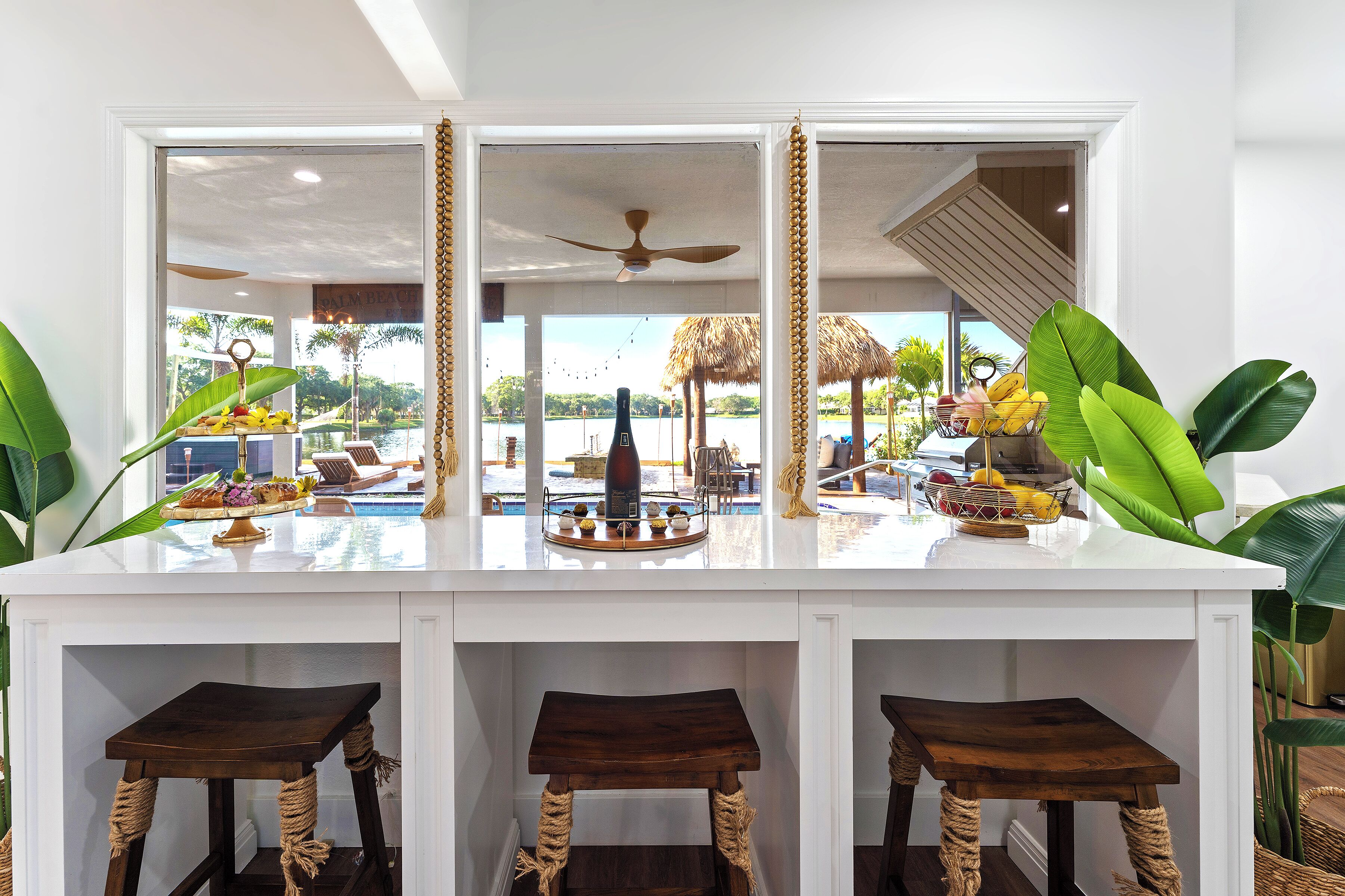 A white bar area with a pool view, wooden stools, and a fruit bowl.