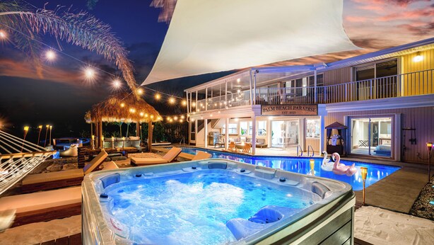 Un jacuzzi en una terraza de madera con sillas reclinables, una piscina con un flotador con forma de cisne, y un edificio con un cartel que dice "Palm Beach Paradise".