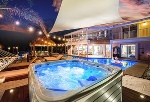 A hot tub on a wooden deck with lounge chairs, a pool with a swan float, and a building with a sign reading "Palm Beach Paradise". - Palm Beach Paradise | Villa w/ Beach, Pool, Cinema & Tiki (Palm Beach Gardens)