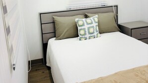 A bedroom with a bed, a bedside table, a nightstand, a wall-mounted air conditioner, and a window with blinds.