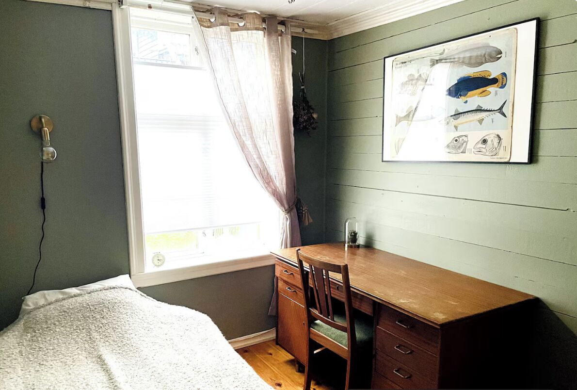 Een slaapkamer met een bed, een houten bureau, een stoel, een raam met gordijnen en een illustratie van een vis in een lijst aan de muur.