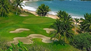 Un parcours de golf offrant une vue sur la plage, bordé de palmiers et avec une chaîne de montagnes en arrière-plan.