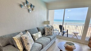 Ein Wohnzimmer mit grauem Sofa, gemusterten Kissen, einem runden Couchtisch und Blick auf den Strand durch große Fenster.