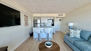 Ein modernes Wohnzimmer mit grauem Sofa, einem Holztisch und einer Küchenzeile mit weißen Schränken und einem Edelstahlkühlschrank.