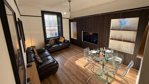 A modern living room with a leather sofa, a glass dining table, and a TV mounted on the wall. - Ayr Town Centre Apartment - 3 Bedrooms (Ayr)