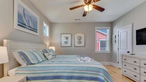 A bedroom with a bed, a ceiling fan, a dresser, and artwork on the walls. - Butterfly Oasis (Carolina Beach)