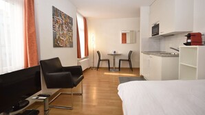A hotel room with a bed, a desk with a computer, a chair, a small table with two chairs, a kitchen area with a microwave, and a painting on the wall.