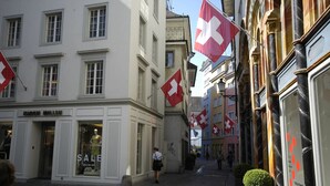 A cobblestone street with buildings featuring traditional architecture, Swiss flags, and a storefront with a sale sign.