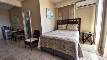 A hotel room with a bed, a desk with a chair, a small table, and an air conditioning unit.