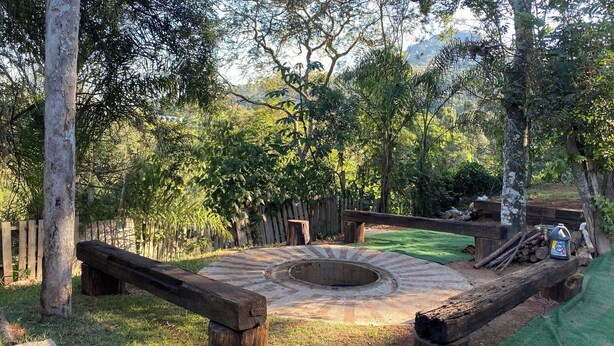 A circular stone fire pit surrounded by a wooden deck, with a wooden fence and trees in the background. - Nature, swimming pool, billiards, barbecue and comfort – Quinta do Remanso (Mairiporã)