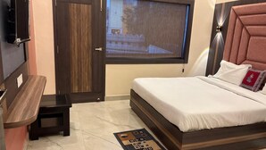 A bedroom with a bed, a small table, a TV mounted on the wall, and a window with a view of the outside. - DM PAYING GUEST HOUSE (VARANASI)