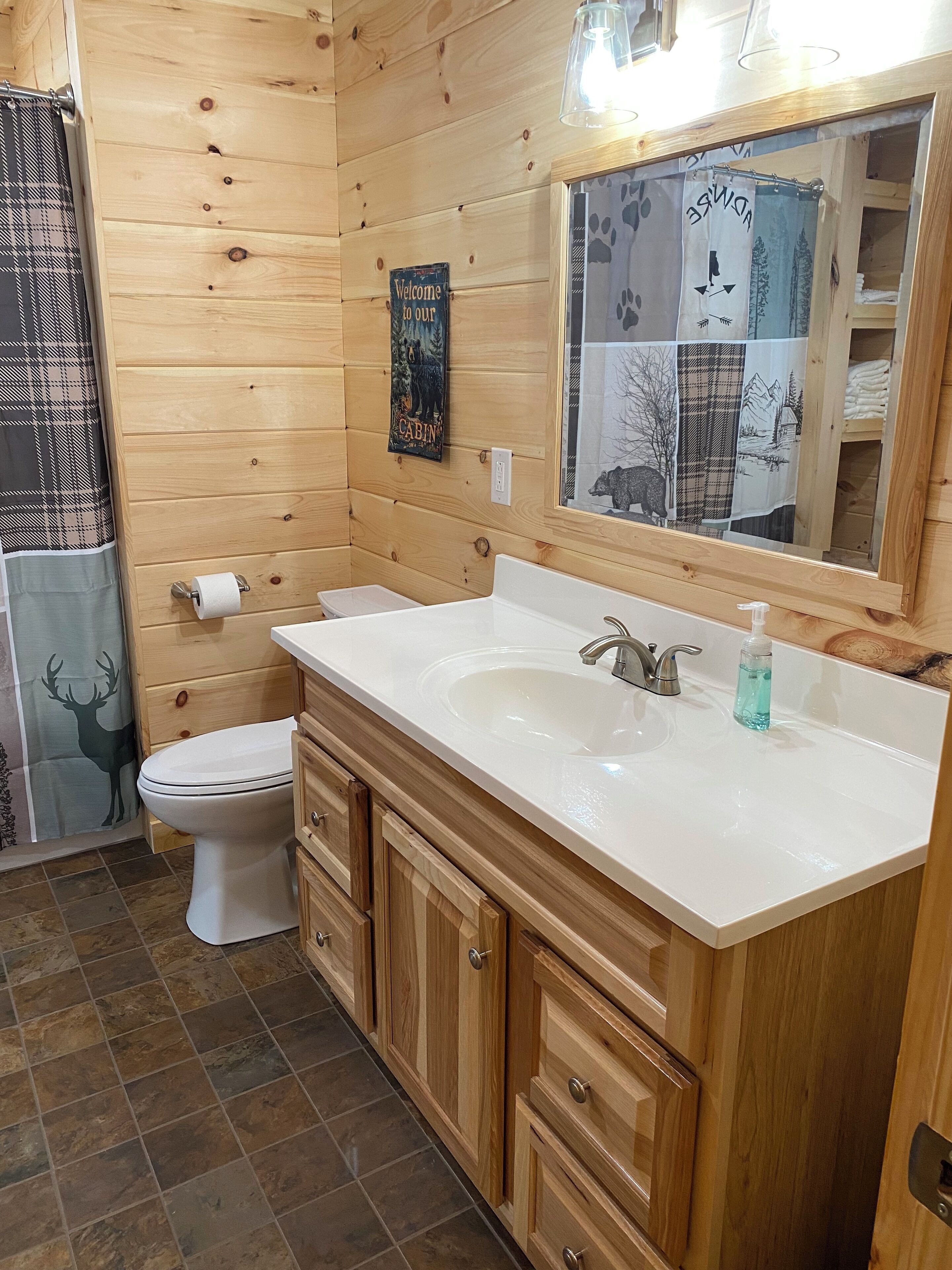 A wooden cabin-style bathroom with a white sink, a toilet, and a mirror with a cabin-themed curtain.