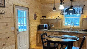 A cozy cabin interior with a wooden interior, a dining area with a table and chairs, a microwave, and a window with a view of trees.