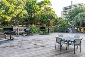 Um pátio externo com deck de madeira, estações de churrasqueira em metal e uma mesa com cadeiras.