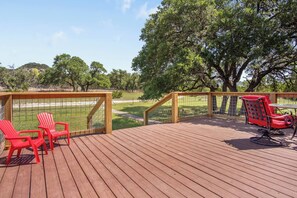 A wooden deck with red chairs and a table, overlooking a grassy area with trees. - Tejas Hills | 4BR Retreat with Firepit & Hill View (Pipe Creek)