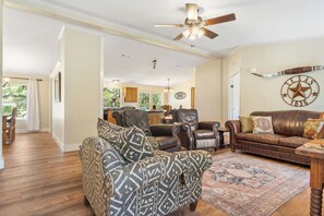 A spacious living room with a large brown leather sofa, patterned armchairs, and a traditional wooden coffee table. A ceiling fan and a decorative wall piece are also visible.