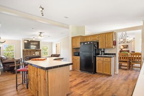 A modern kitchen with wooden cabinets, a black refrigerator, and a dining area with a wooden table and chairs. - Tejas Hills | 4BR Retreat with Firepit & Hill View (Pipe Creek)